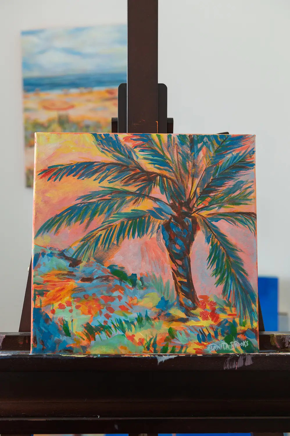 Original palm tree sunset painting titled "Sorbet Skies" by Danita Brooks, framed in white plein air frame shown here on an easel and displayed in natural light
