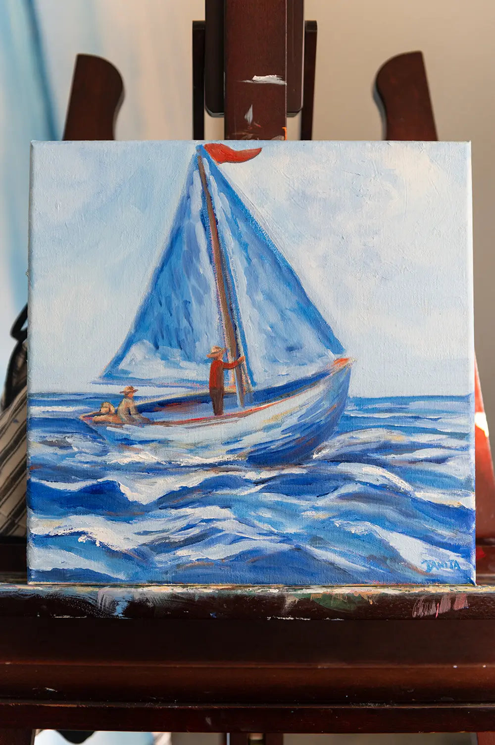 Along for the Ride by Danita Brooks on a wooden easel, depicting 2 figures and dog in a blue-sailed boat on rolling waves.
