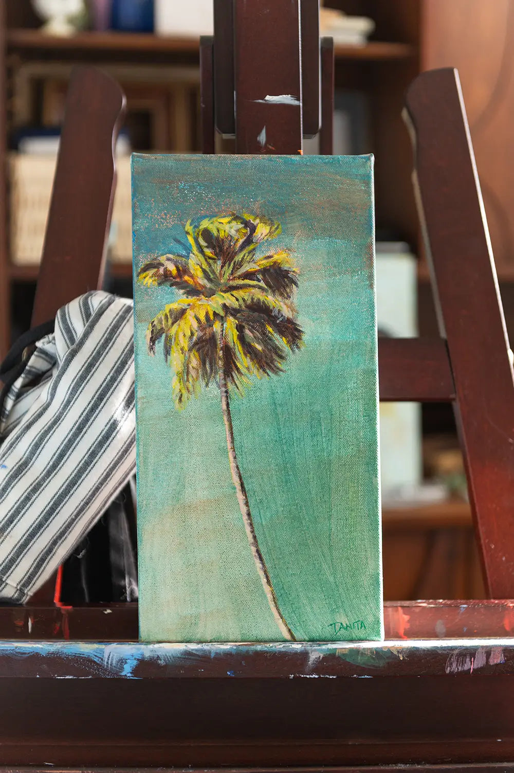 "The Long Way Home" palm tree painting by Danita Brooks on a wooden easel, an acrylic painting of a slender palm with golden-green fronds against a turquoise background.