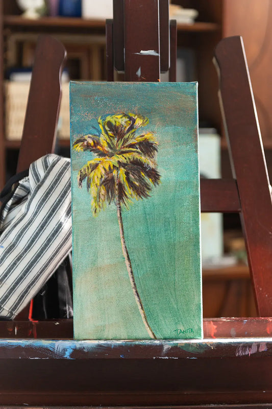 "The Long Way Home" palm tree painting by Danita Brooks on a wooden easel, an acrylic painting of a slender palm with golden-green fronds against a turquoise background.