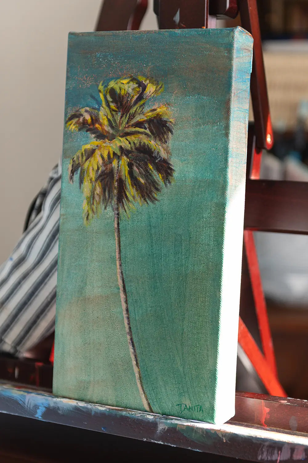 Angled view of "The Long Way Home" on an easel, showing the palm tree painting and the right edge of the stretched canvas.