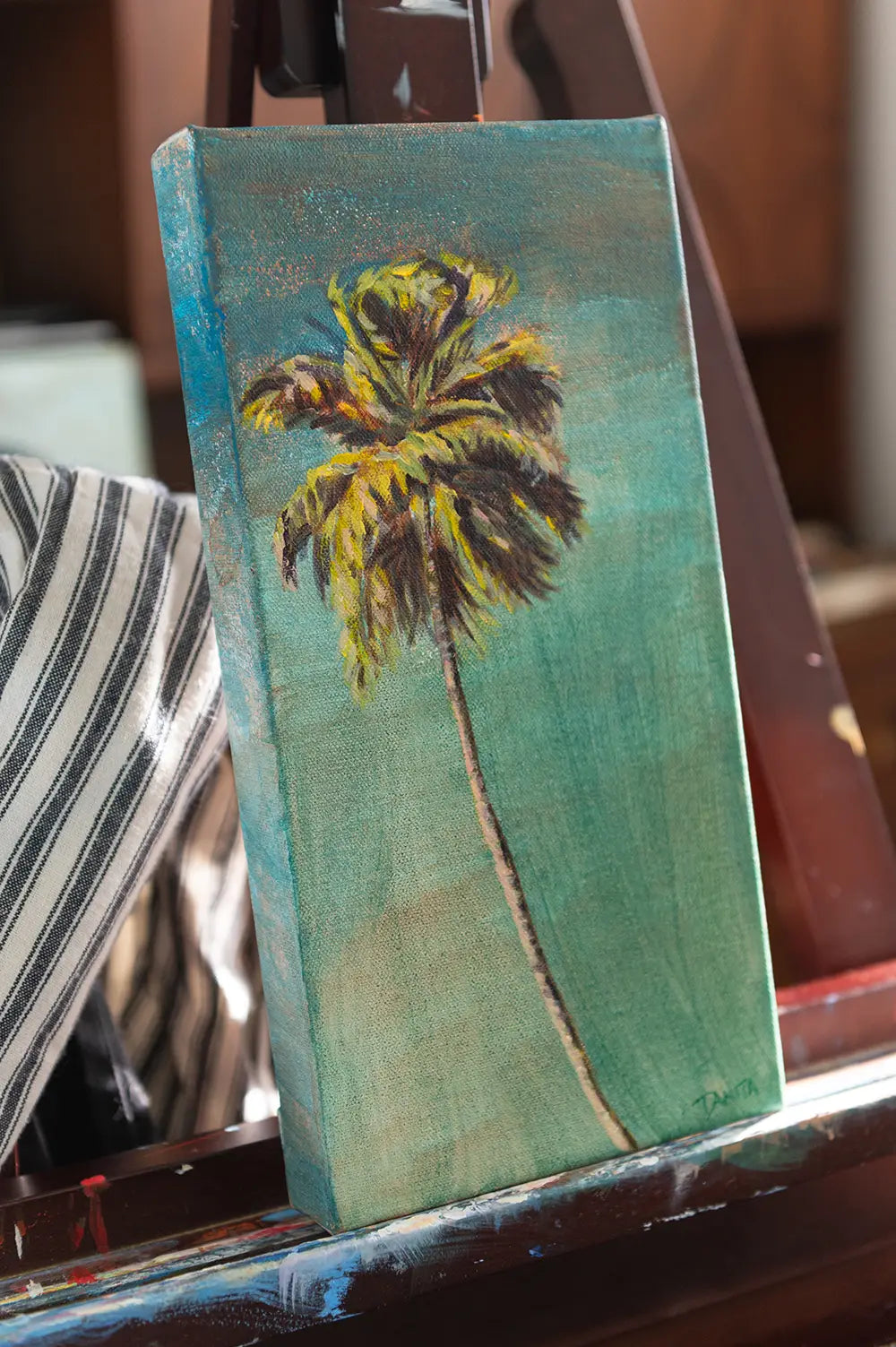 "The Long Way Home" viewed from the opposite angle on a studio easel, highlighting the curved palm trunk and textured turquoise background.