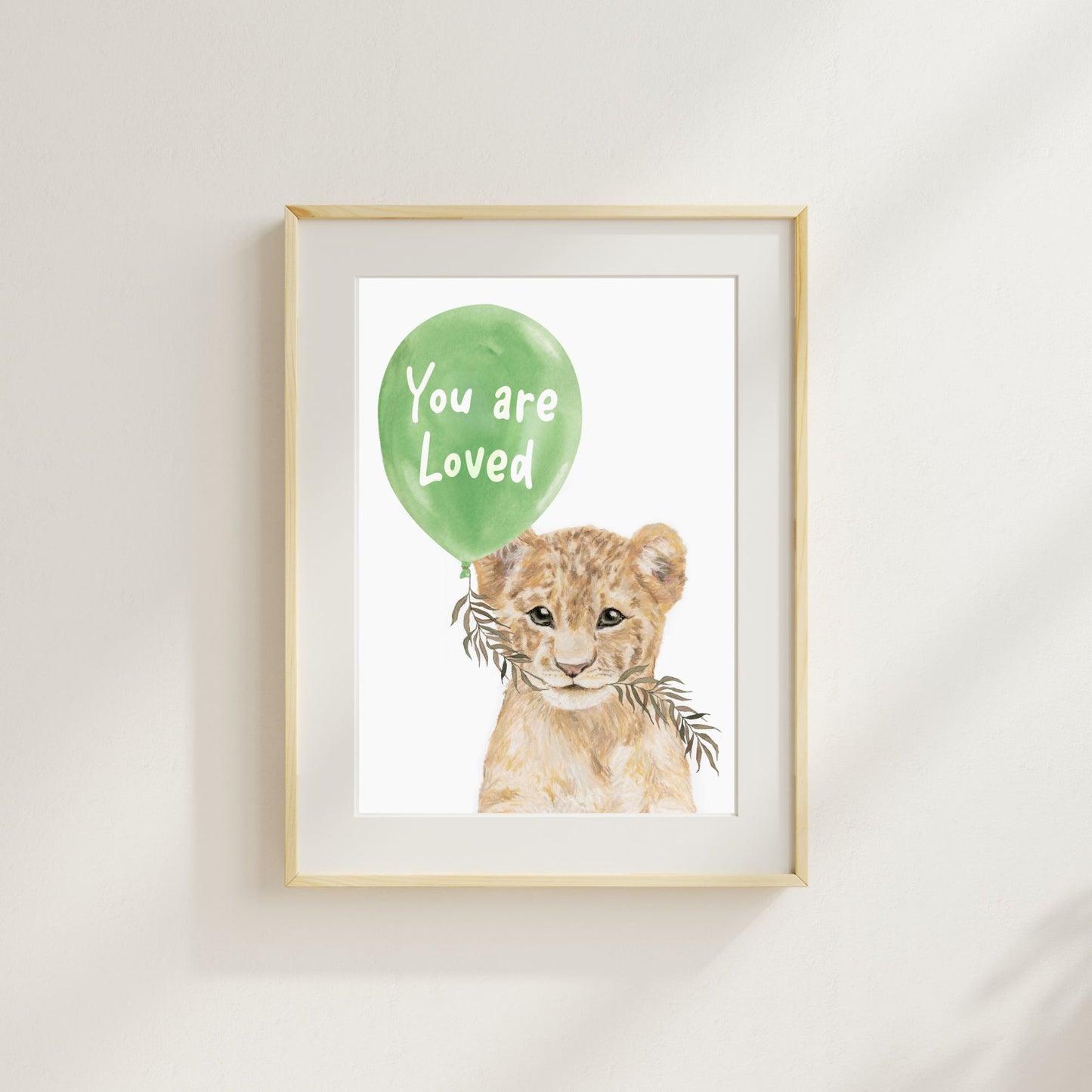 Framed artwork of a lion cub with a green balloon that says 'You are Loved' on a white wall.
