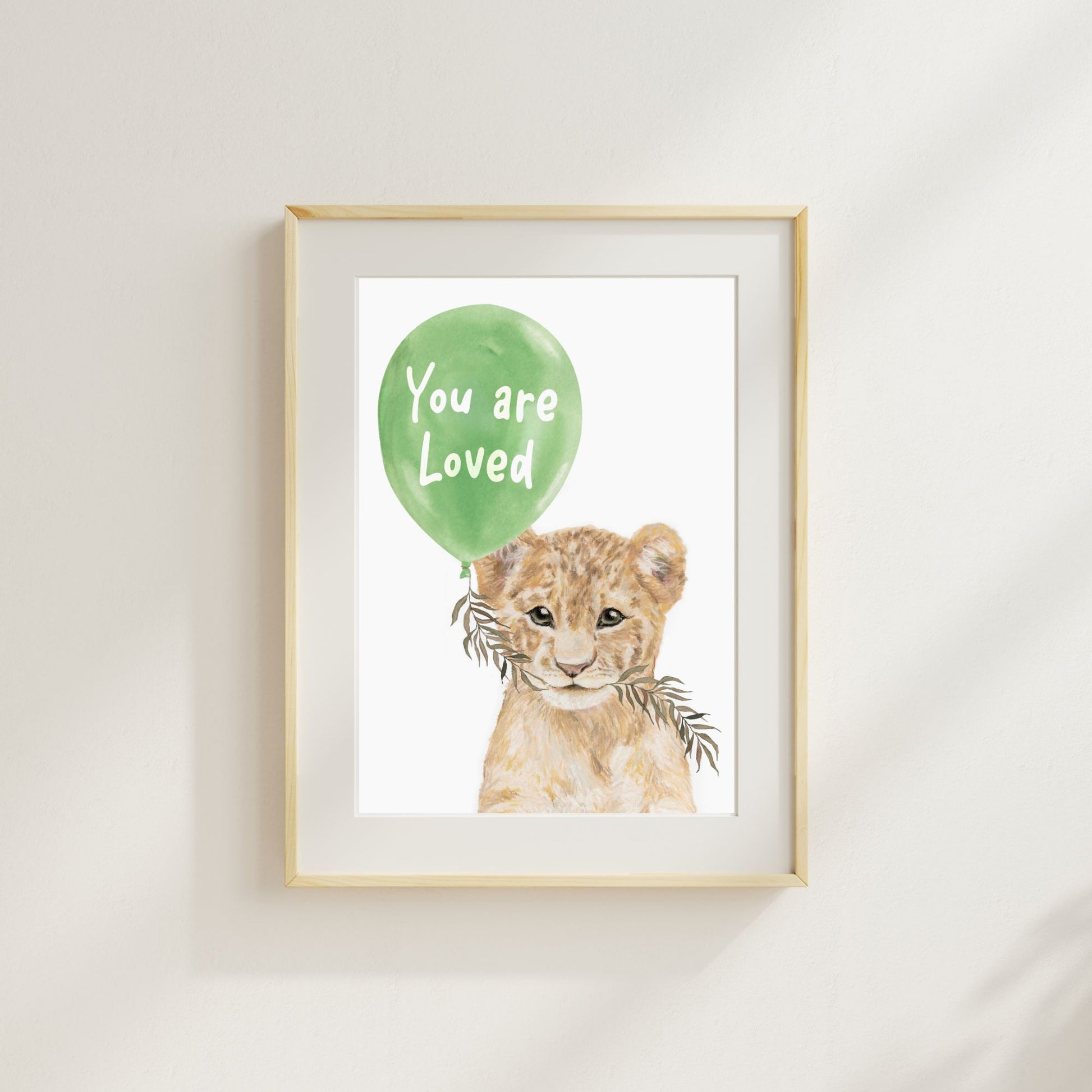 Framed artwork of a lion cub with a green balloon that says 'You are Loved' on a white wall.